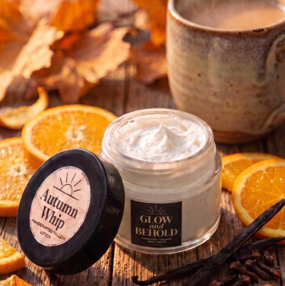 A jar of Glow Before Behold face and body tallow lotion with its lid off, placed on a rustic wooden table surrounded by orange slices, cinnamon sticks, cloves, dried leaves, and a steaming mug.