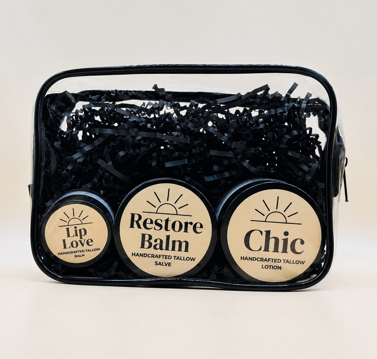 A clear zippered cosmetic pouch filled with black shredded paper and three round tins of handcrafted tallow salves  and lotion labeled Lip Love, Restore Balm, and Chic, on a light beige background.