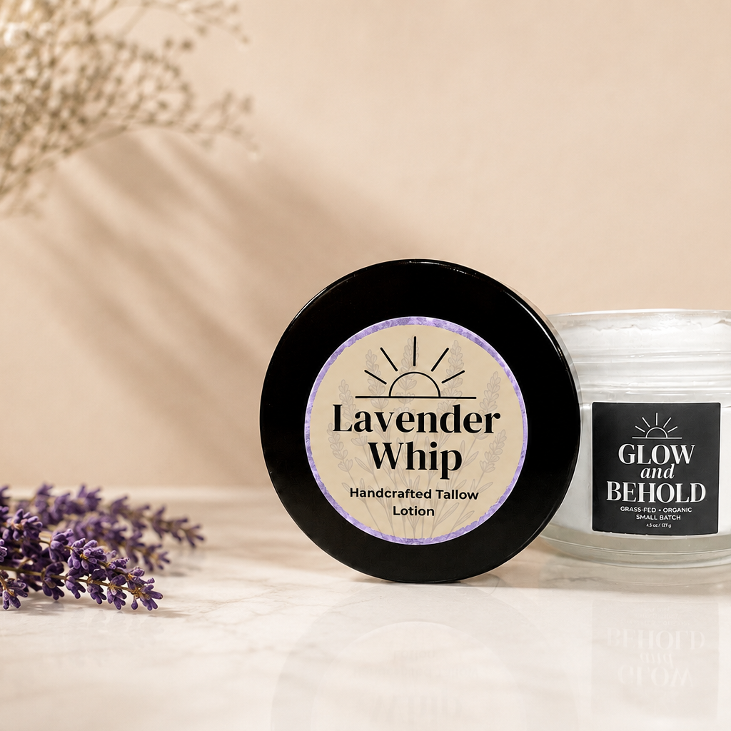A jar of Glow and Behold Handcrafted Tallow Lotion in lavender scent with its lid placed beside it, styled on a marble surface with dried lavender, a beige vase, and a folded towel against a warm beige background.
