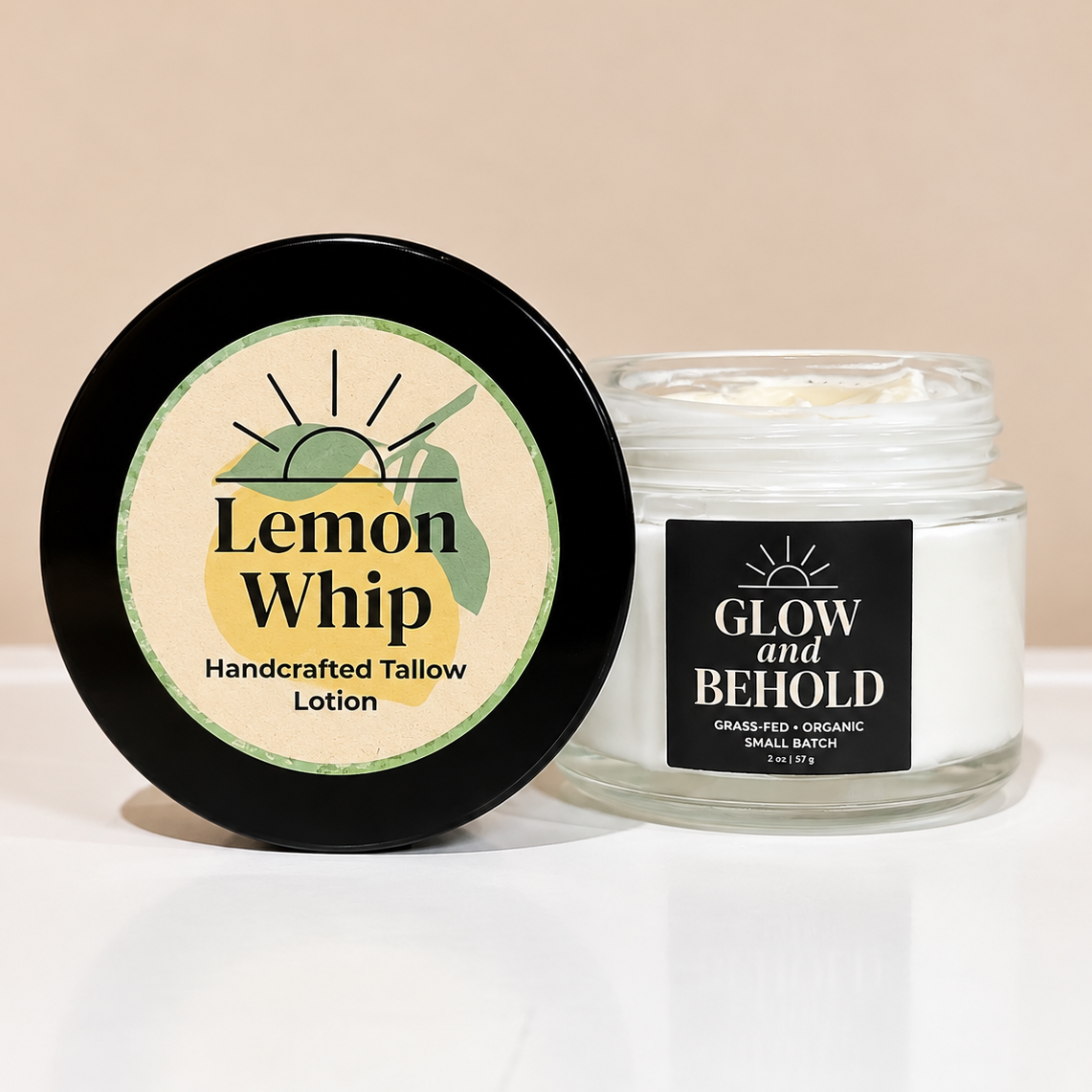 A jar of Lemon Whip Handcrafted Tallow Lotion by Glow and Behold, shown with its black lid placed in front of the clear glass jar containing white tallow lotion.