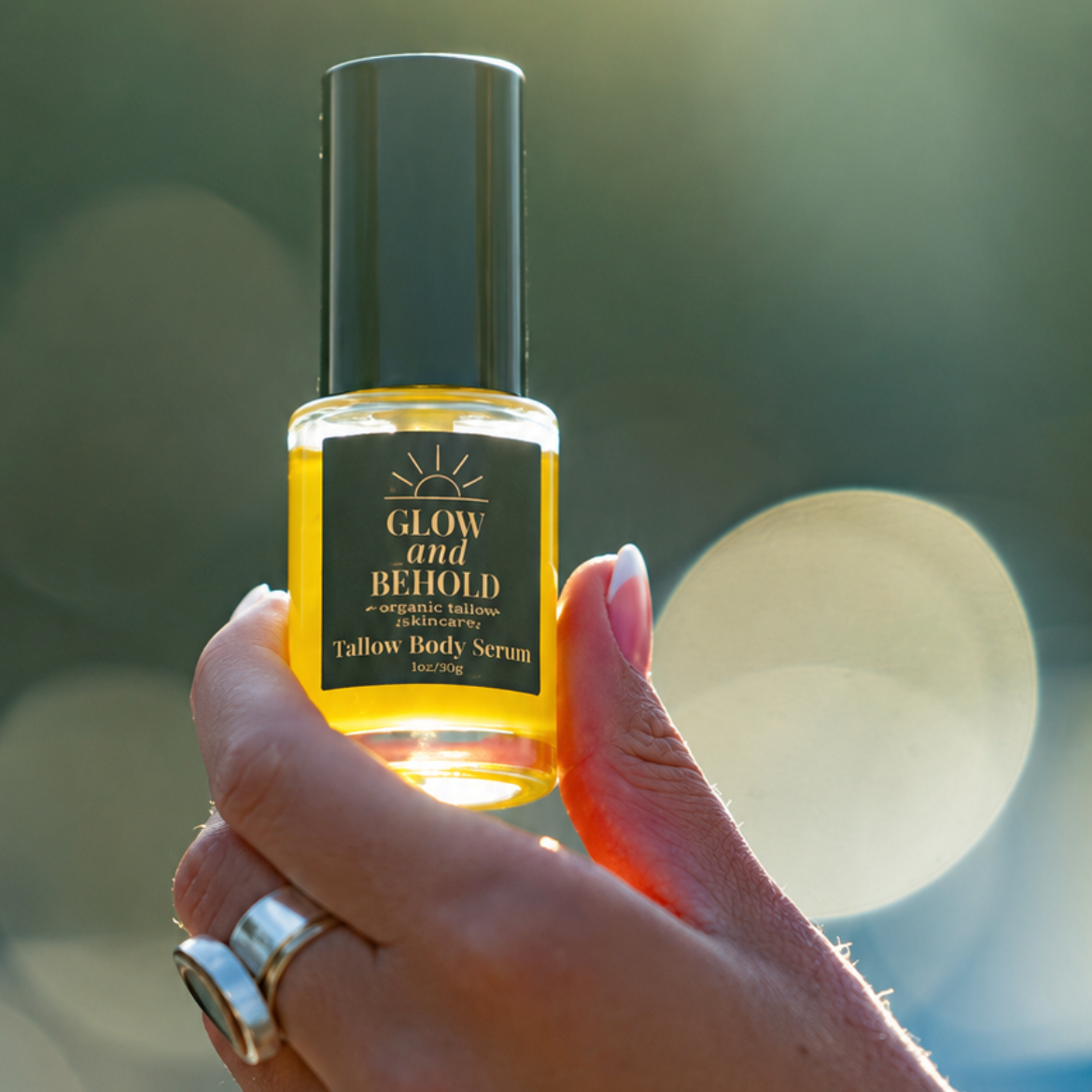 A hand holding a small glass bottle of Glow and Behold Tallow Body serum oil against a soft, blurred outdoor  background