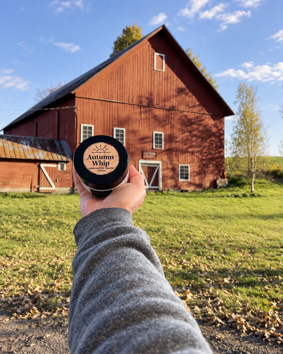 Glow & Behold Autumn Whip tallow lotion held in front of a red barn in a fall countryside setting